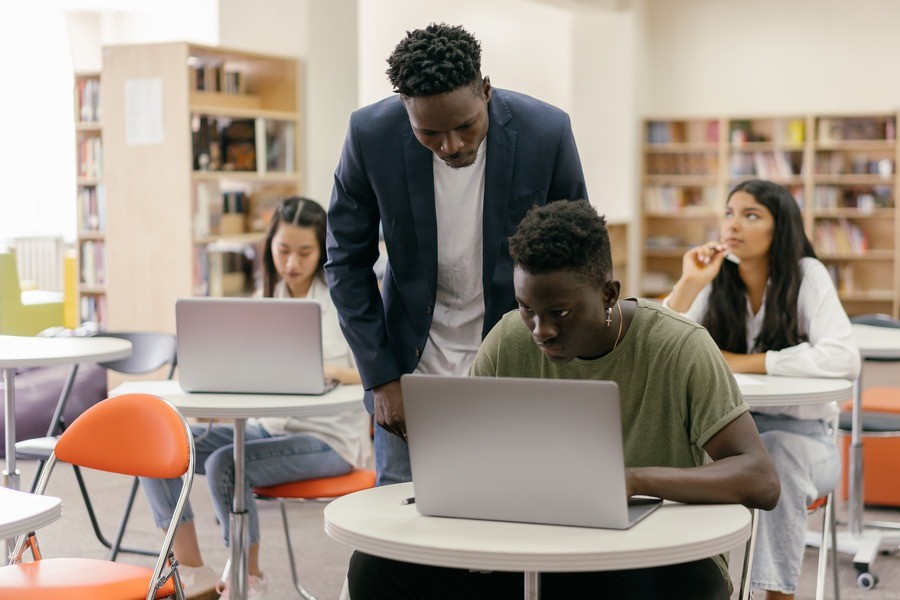 Professor wearing a blazer leaning over a students shoulder checking the work he’s doing on the laptop, which sits open on his desk.