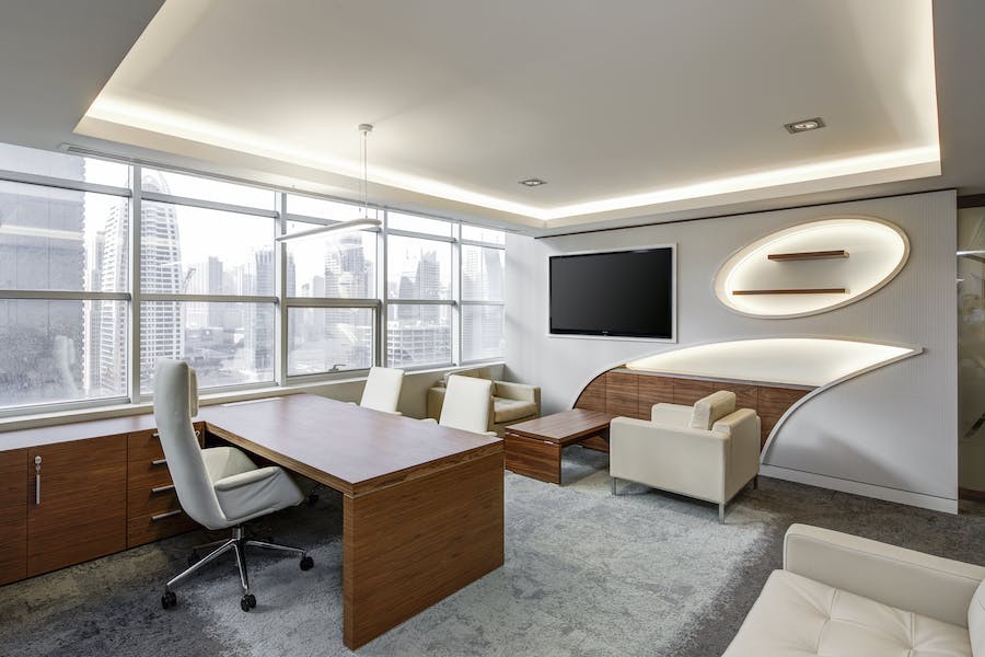 A modern office with desks, chairs, large windows, and a television mounted on the wall.