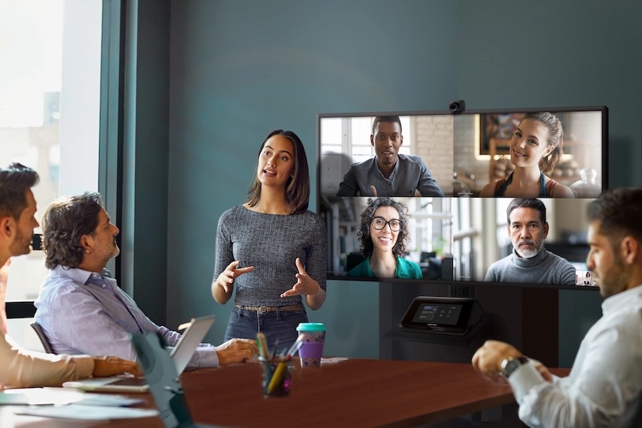 A woman leading a hybrid meeting, with three people in-persona t a table and four people joining remote on a TV screen.