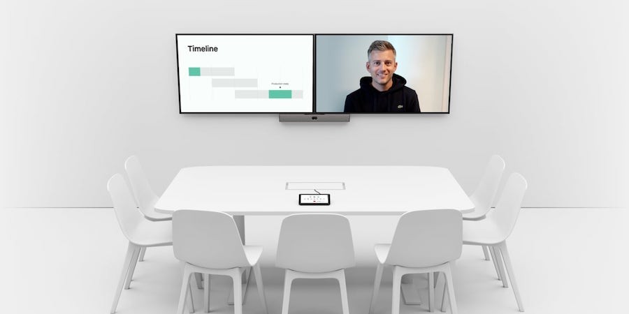 A Tampa office video conferencing system with dual screens showing a timeline and video call participant above a white table.