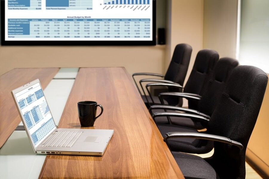 A meeting room showing a seat and laptop on the table with a large screen display in the background.